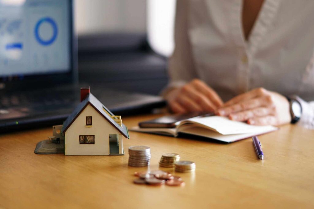 Immobilienbewertung und Finanzen – Hausmodell mit Geldmünzen auf Tisch, Makler analysiert Marktpreise